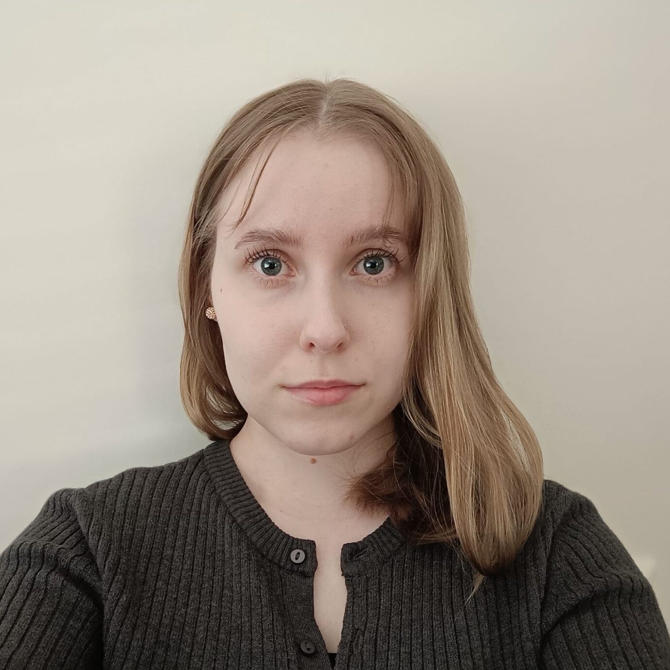 Nuori nainen, jolla on pitkät vaaleanruskeat hiukset ja vaalea iho, katsoo suoraan kameraan. Hänellä on päällään tumma villapaita ja hän seisoo vaalean taustan edessä.