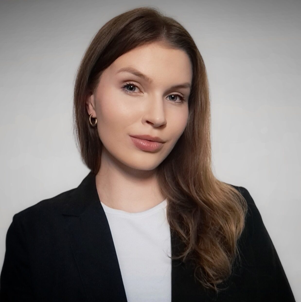 Vaaleaihoinen nainen, jolla on pitkät, vaaleanruskeat hiukset, pukeutuu mustaan bleiseriin valkoisen topin päällä. Hän katsoo kameraan neutraali ilme kasvoillaan vaaleanharmaata taustaa vasten.