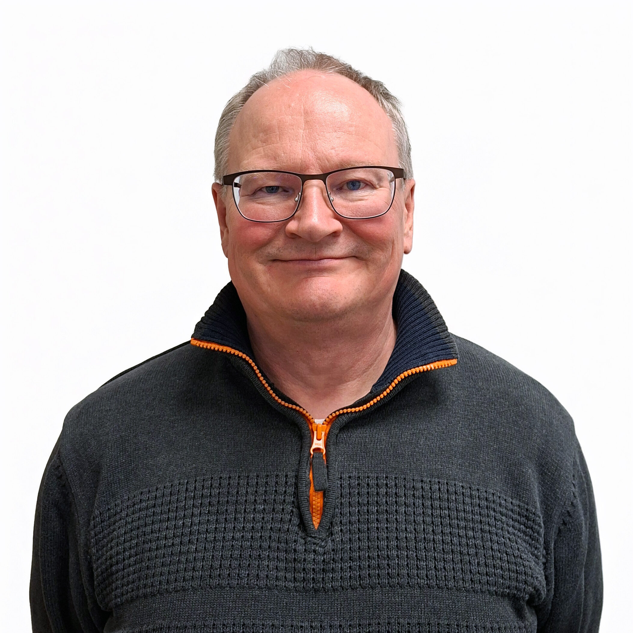 Keski-ikäinen mies, jolla on lyhyet harmaat hiukset ja silmälasit, hymyilee ja jolla on yllään tumma neulepusero, jossa on oranssi vetoketju ja kaulus, ja joka seisoo valkoista taustaa vasten.