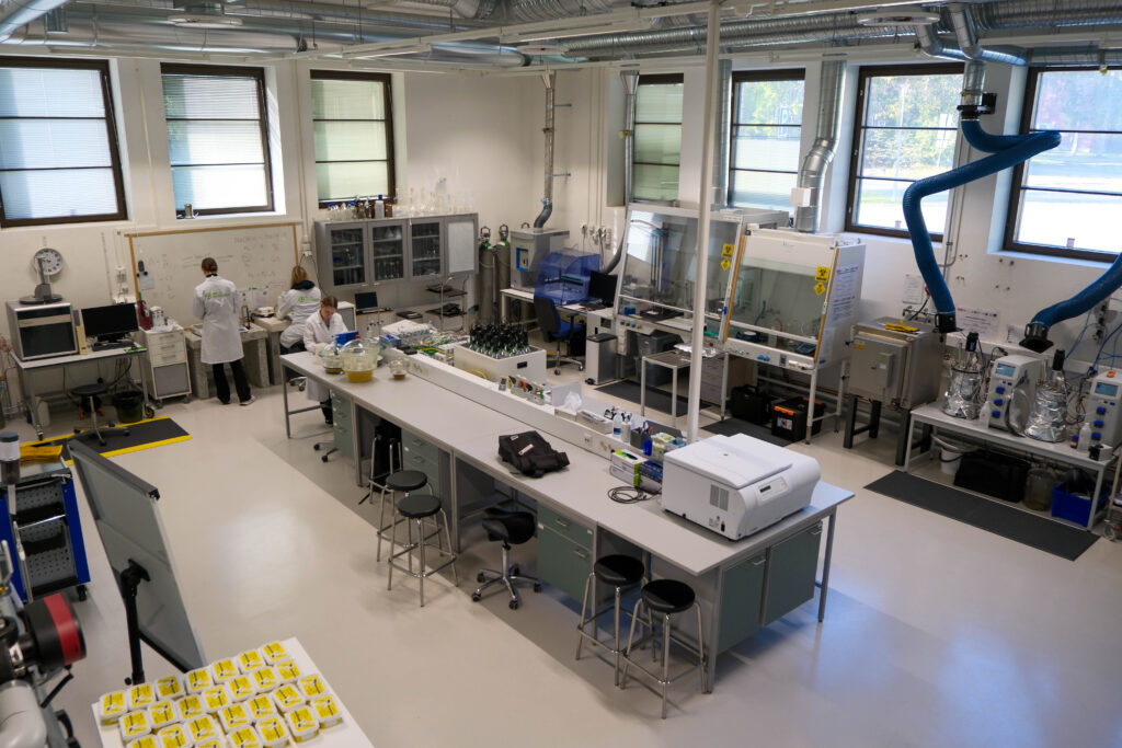Nykyaikainen laboratorio, jossa valkotakkiset tutkijat työskentelevät; laboratoriopenkit ovat täynnä laitteita, tietokoneita, instrumentteja ja tarvikkeita. Suuret ikkunat tuovat luonnonvaloa, ja katosta roikkuu sinisiä ilmanvaihtovarsia.