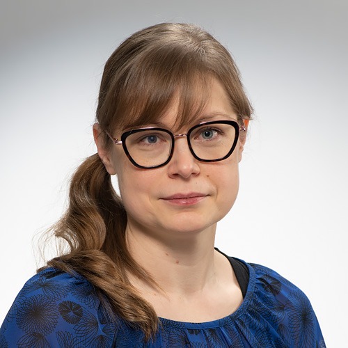 Nainen, jolla on vaaleanruskeat hiukset sivuponinhännässä, suuret mustat silmälasit ja sinikuvioinen toppi, katsoo kameraan neutraalilla ilmeellä vaaleaa taustaa vasten.