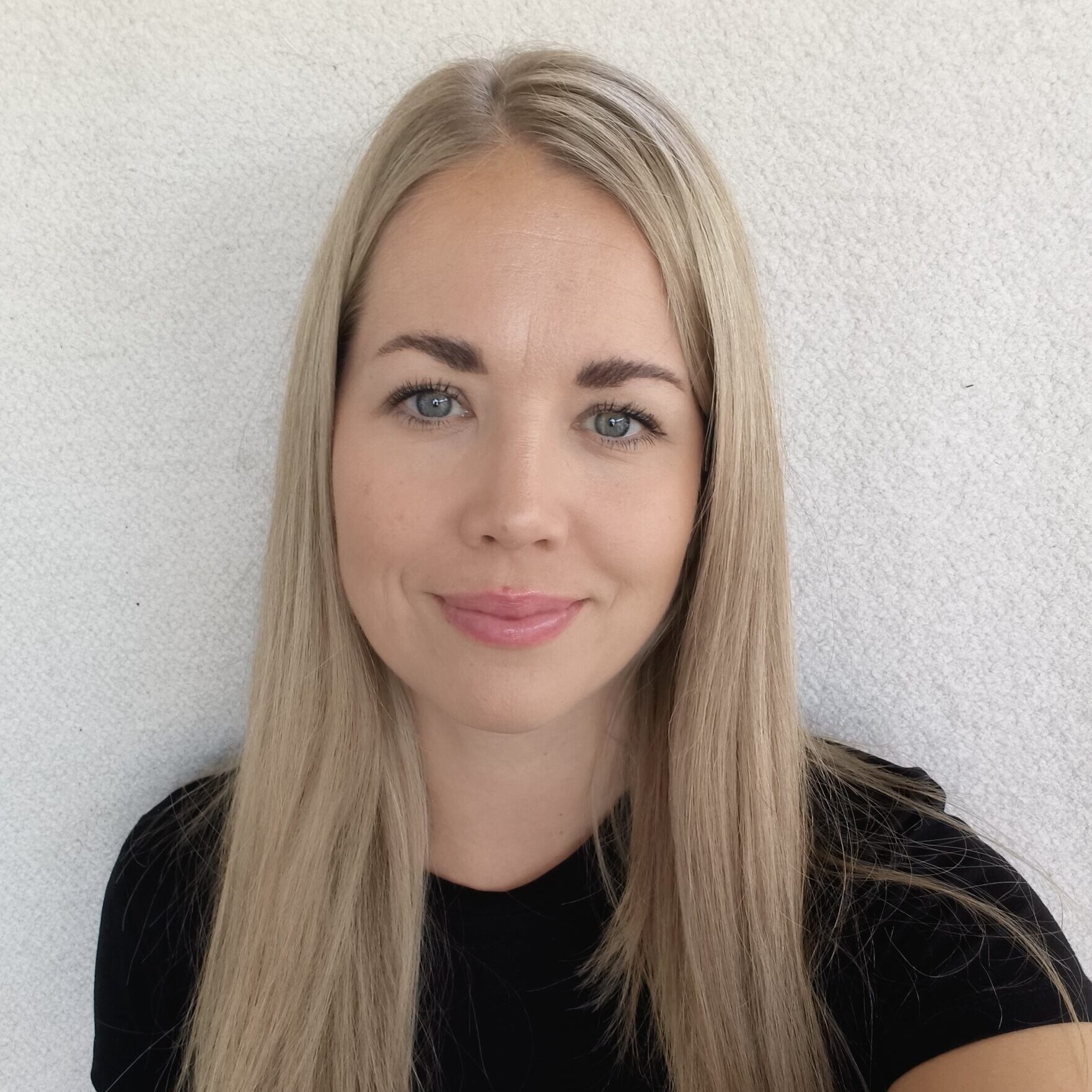 Nainen, jolla on pitkät suorat vaaleat hiukset ja siniset silmät, hymyilee lempeästi kameralle. Hänellä on yllään musta toppi ja hän seisoo tavallisen, vaalean taustan edessä.