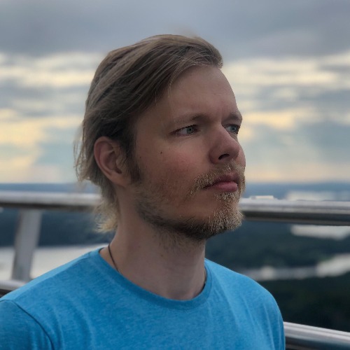 Vaaleat hiukset ja lyhyt partainen mies, jolla on sininen paita, seisoo ulkona metallikaiteen edessä, jonka taustalla on maisemallinen tausta, jossa on puita, vettä ja pilvinen taivas auringonlaskun aikaan.