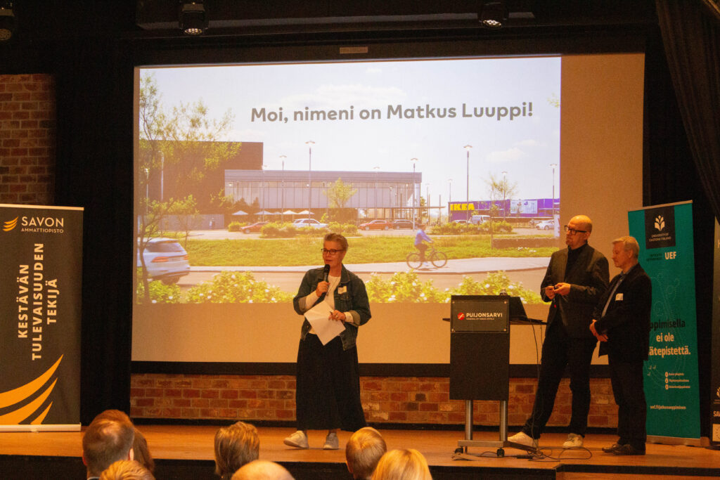 Kolme ihmistä seisoo lavalla esittelemässä yleisölle. Heidän takanaan olevalla ruudulla näkyy suomeksi Moi, nimeni on Matkus Luuppi! ja ulkokuvassa on tavaratalo ja pyöräilijä.