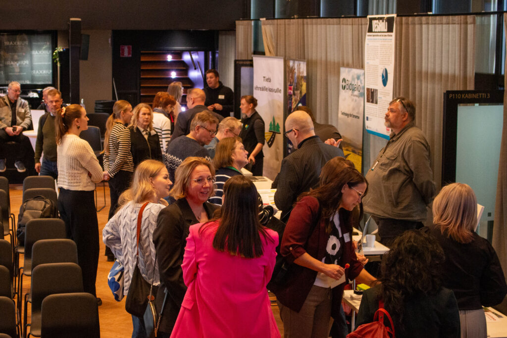 Ryhmä ihmisiä on vuorovaikutuksessa ja verkostoituu näyttelypöytien ympärillä sisätiloissa järjestettävässä tapahtumassa, ja taustalla näkyy tiedotusjulisteita ja banderolleja. Jotkut osallistujat keskustelevat, kun taas toiset tarkkailevat näyttelyitä.