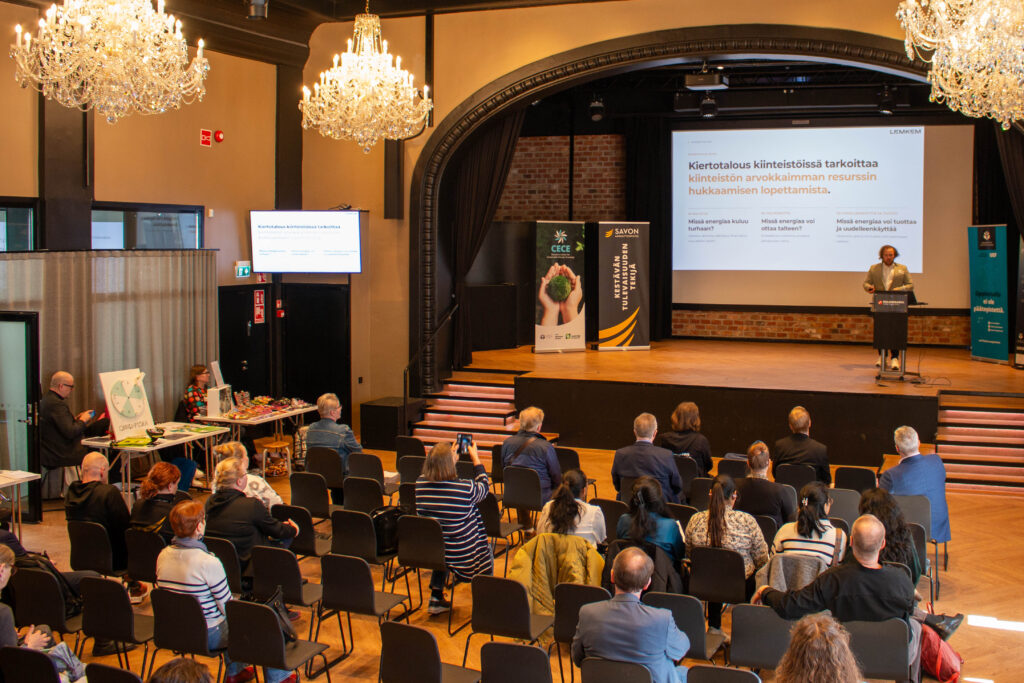 Puhuja esitelmöi konferenssissa hyvin valaistussa salissa, jossa on kattokruunut, ja osallistujat kuuntelevat. Suurella näytöllä näytetään dioja, ja sivulle on pystytetty pöytiä, joissa on materiaalia.