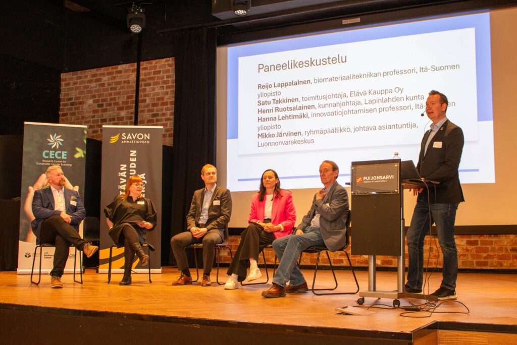 Kuusi liikemiespukuista henkilöä osallistuu lavalla käytävään paneelikeskusteluun. Viisi istuu, kun taas yksi seisoo ja puhuu korokkeella. Heidän takanaan olevalla näytöllä näkyy tietoa paneelista, ja vasemmalla näkyy bannereita.