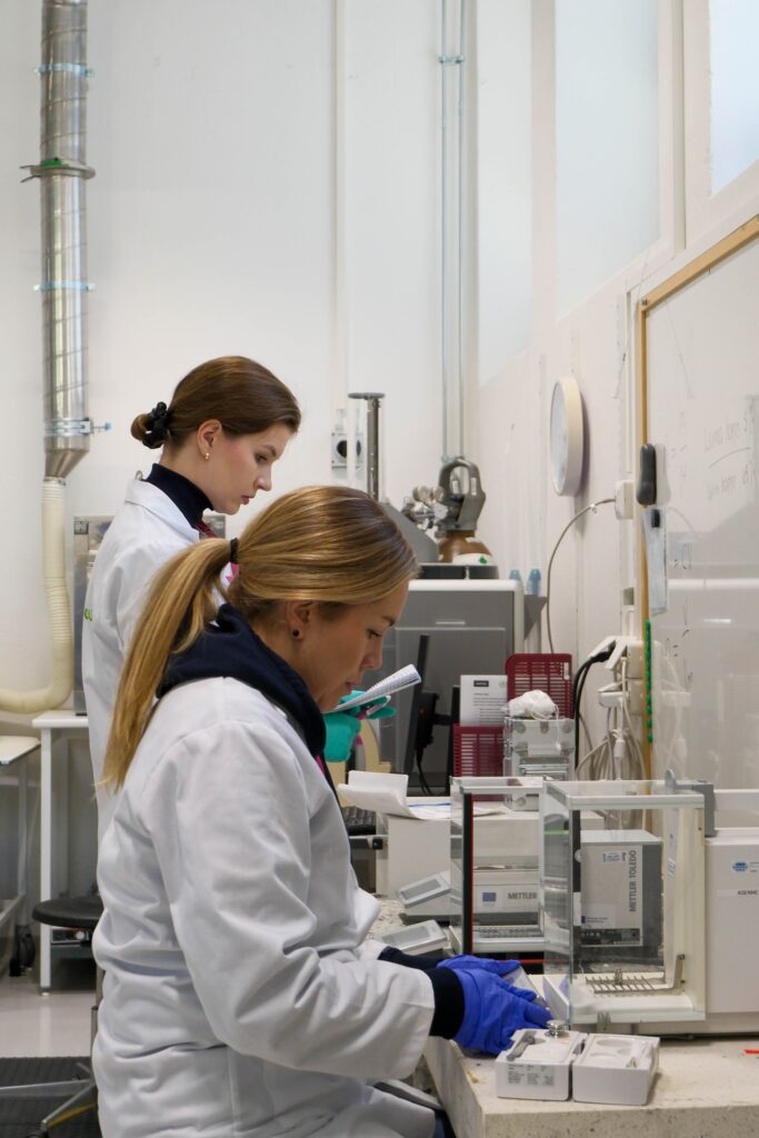 Kaksi laboratoriotakkiin pukeutunutta naista työskentelee laboratoriotyöpisteessä tieteellisten laitteiden kanssa, käsittelee näytteitä ja kirjaa tietoja. Taustalla on laboratoriolaitteita, hyllyjä ja valkotaulu.