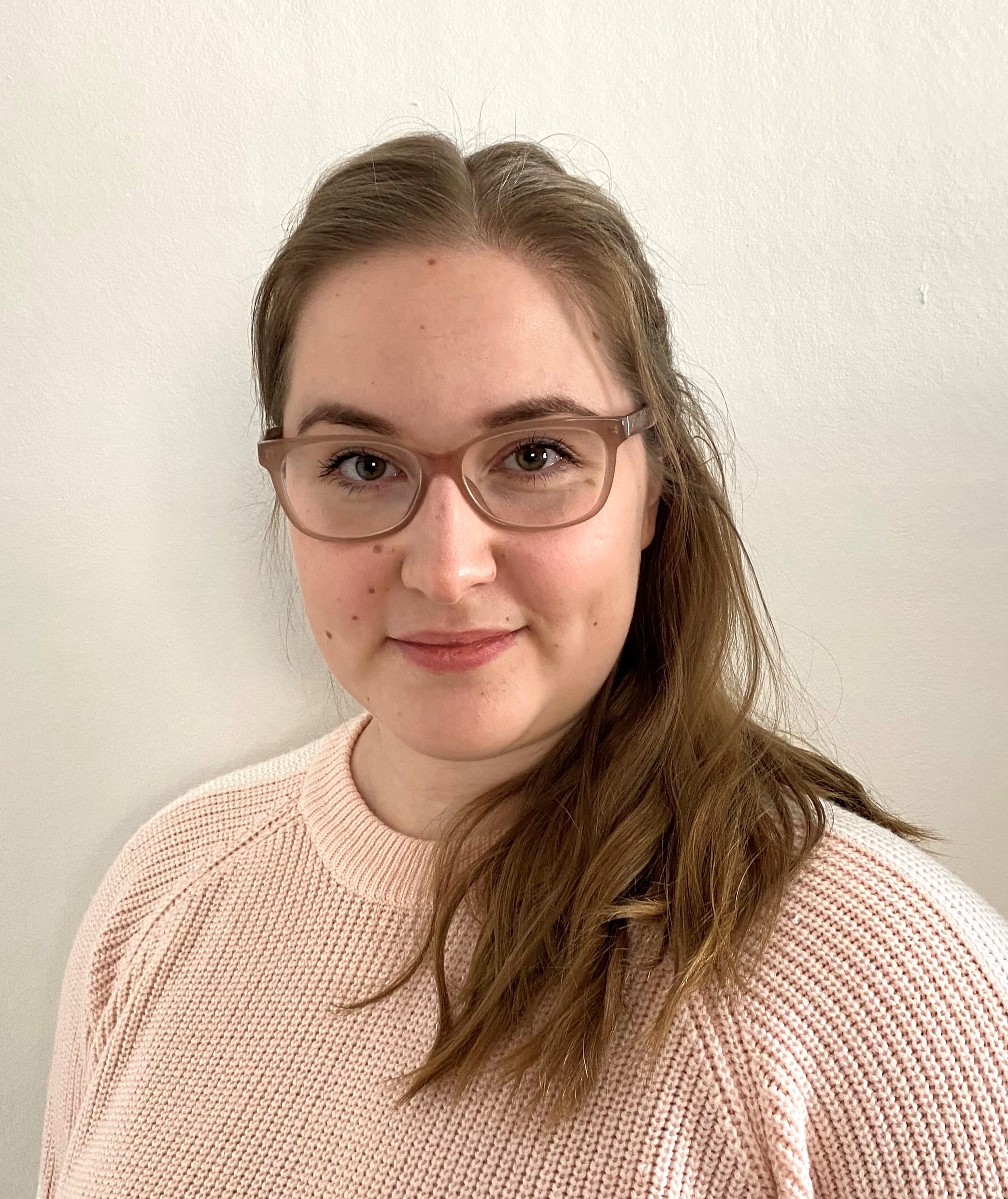 Nainen, jolla on pitkät, vaaleanruskeat hiukset ja silmälasit, hymyilee hieman. Hänellä on yllään vaaleanpunainen neulepusero ja hän seisoo valkoista taustaa vasten.