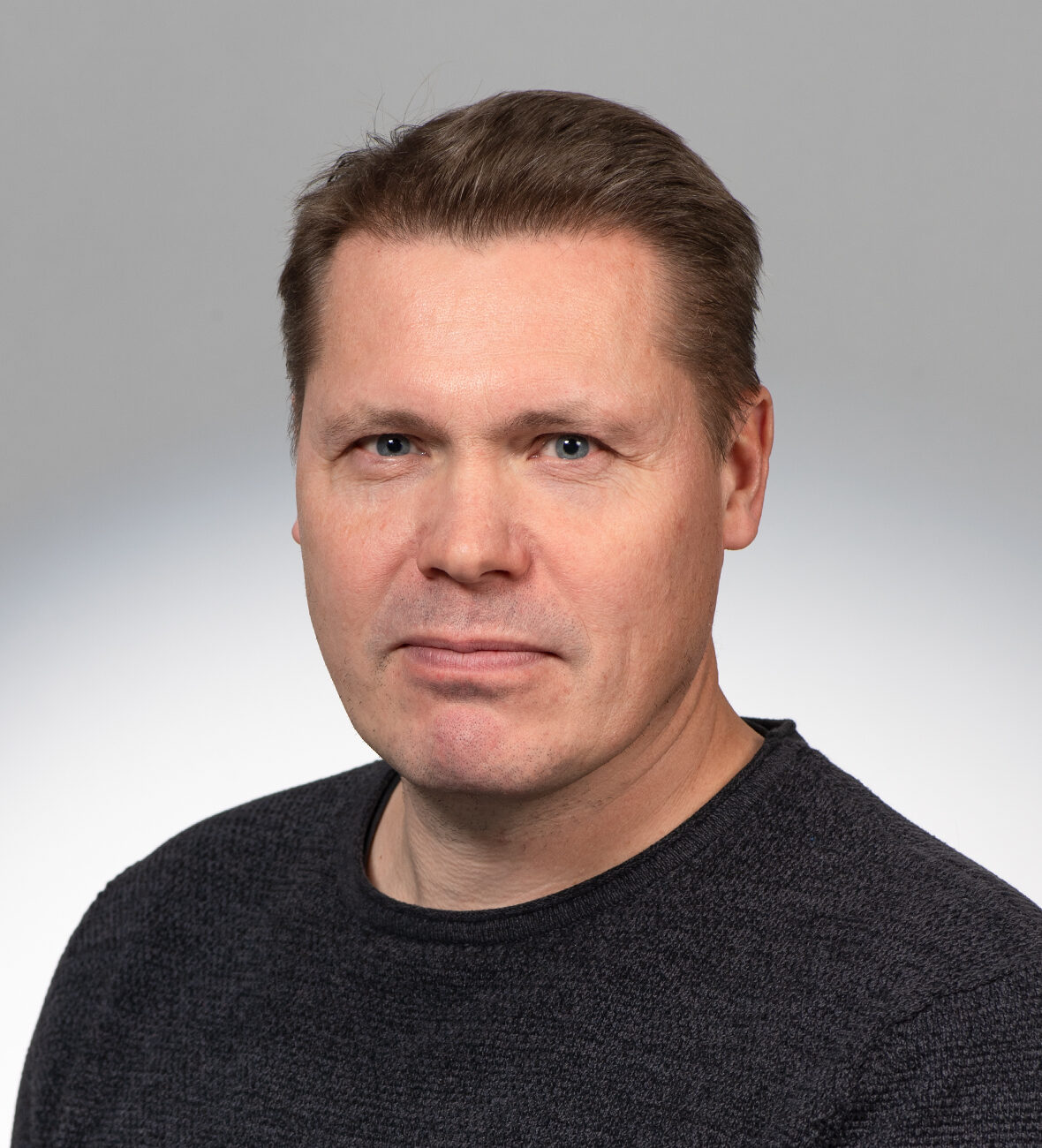 Keski-ikäinen mies, jolla on lyhyet vaaleanruskeat hiukset ja tumma pusero, poseeraa vaaleanharmaan taustan edessä. Hän katsoo suoraan kameraan neutraalilla ilmeellä.
