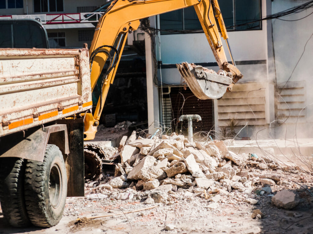 Keltainen kaivinkone siirtää raunioita kuorma-auton vieressä purkutyömaalla; paikalla näkyy betoniraunioita ja hajallaan olevia metallitankoja.