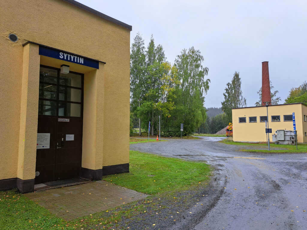 Kivitiellä seisoo keltainen rakennus, jonka kyltissä lukee ”SYTYTIN”, ja sitä ympäröivät puut ja vehreys. Taustalla pilvisen taivaan alla näkyy toinen keltainen rakennus, jossa on korkea savupiippu.