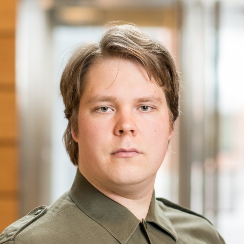 Vaaleaihoinen henkilö, jolla on lyhyet, vaaleanruskeat hiukset ja joka on pukeutunut oliivinvihreään kauluspaitaan, seisoo sisätiloissa ja katsoo suoraan kameraan ilmeettömästi.